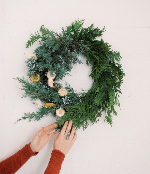 evergreen wreath with natural decorations held against white wall