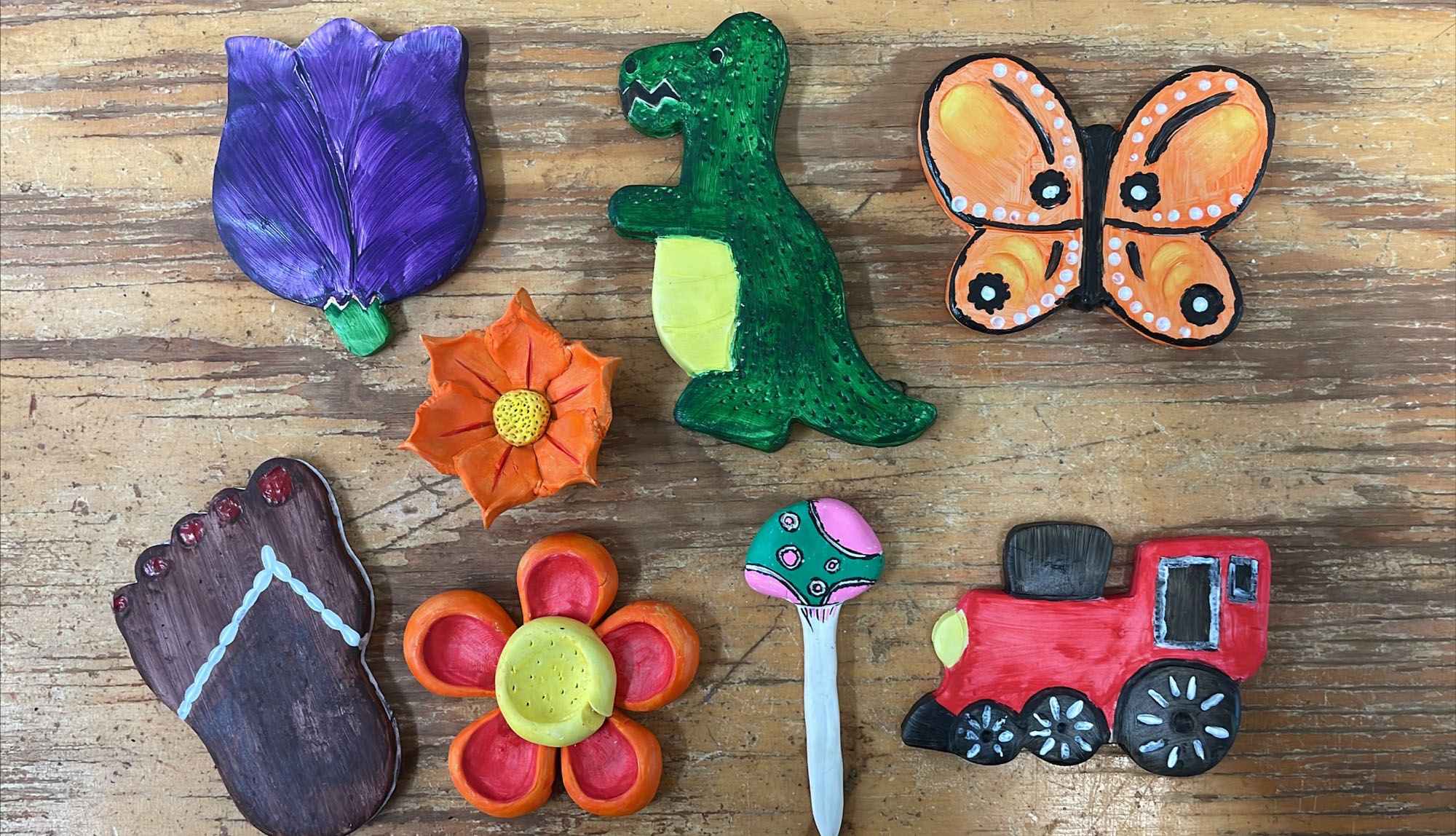 Examples of finished ceramic pieces brightly painted in shapes of flowers, butterflies, dinosaur and mushroom