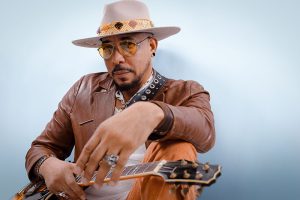 Musician/singer Charlie Redd wearing hat, brown jacket, holding guitar